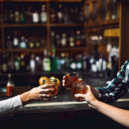 People clinking glasses filled with drinks at a bar, surrounded by shelves stocked with various bottles in the background.