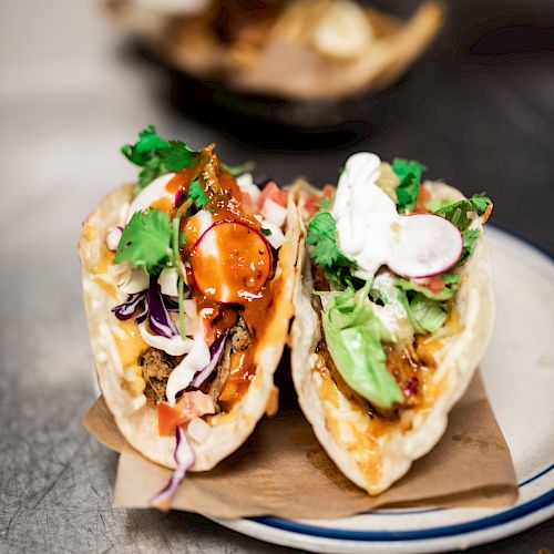 The image shows two tacos on a white plate with various toppings, including herbs, sliced radishes, and cabbage, set on a piece of brown paper.