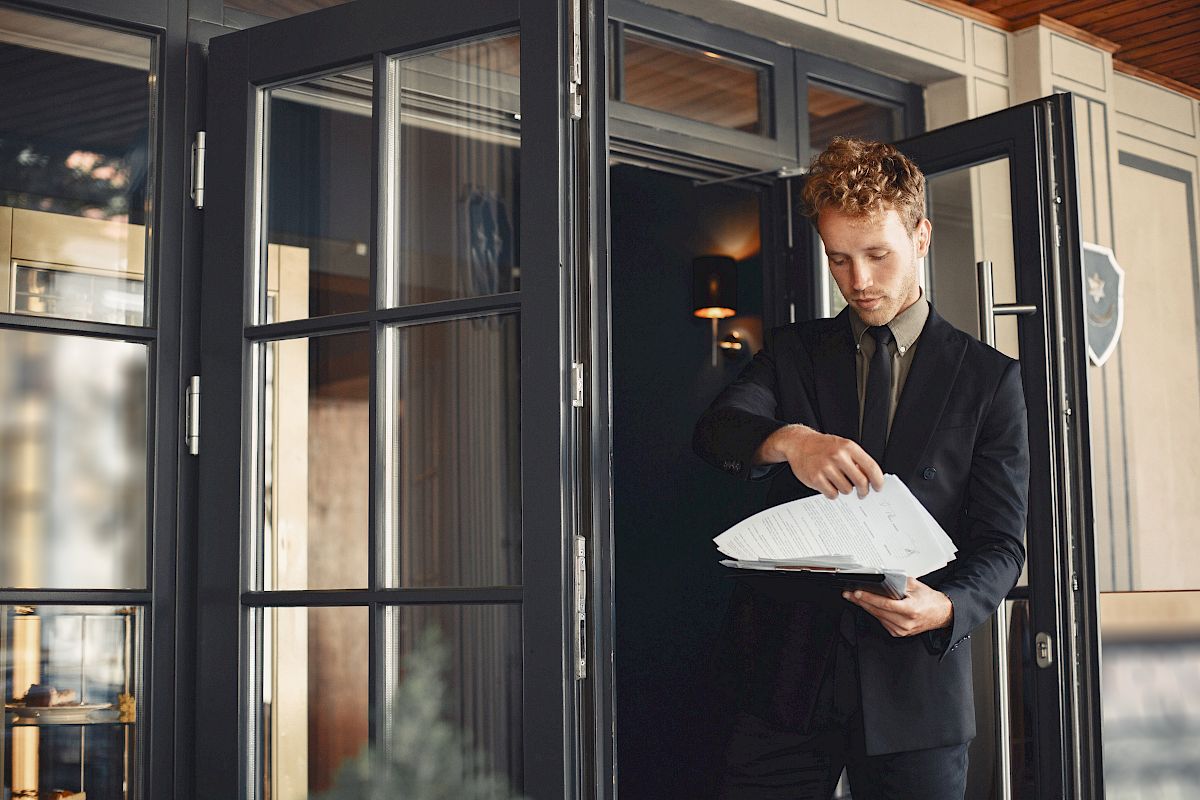 A man in a suit stands outside a building with open glass doors, looking at a stack of papers or files in his hands.