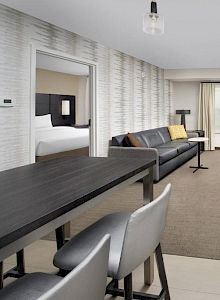 The image shows a modern hotel suite with a living area, desk, and chairs, along with two beds in separate rooms within one suite, ending the sentence.