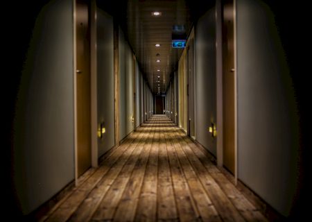 A long, dimly lit hallway with wooden flooring and doors on both sides, leading to darkness in the distance, creates a mysterious ambiance.