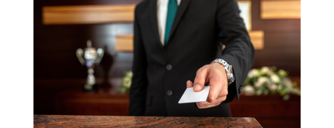 A person in a suit, standing behind a desk or counter, is handing over a card. The background includes a trophy and flowers arranged on a table.