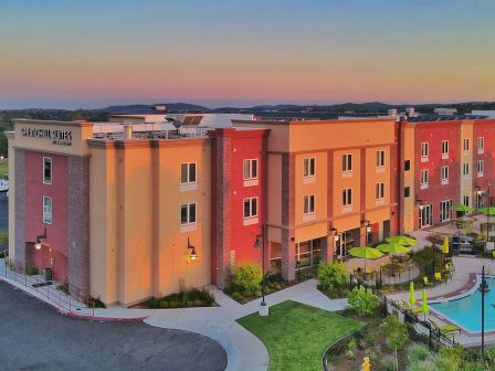 The image shows a multi-story hotel complex at sunset, with a vibrant exterior, a pool, and outdoor seating area surrounded by landscaped greenery.