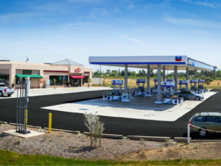 The image shows a gas station with multiple fueling pumps, a convenience store nearby, and several cars parked around the area.