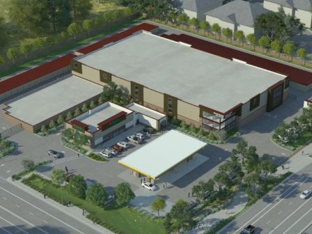 An aerial view of a building complex with a central large structure, surrounding smaller buildings, and a gas station at the entrance, near a busy road.