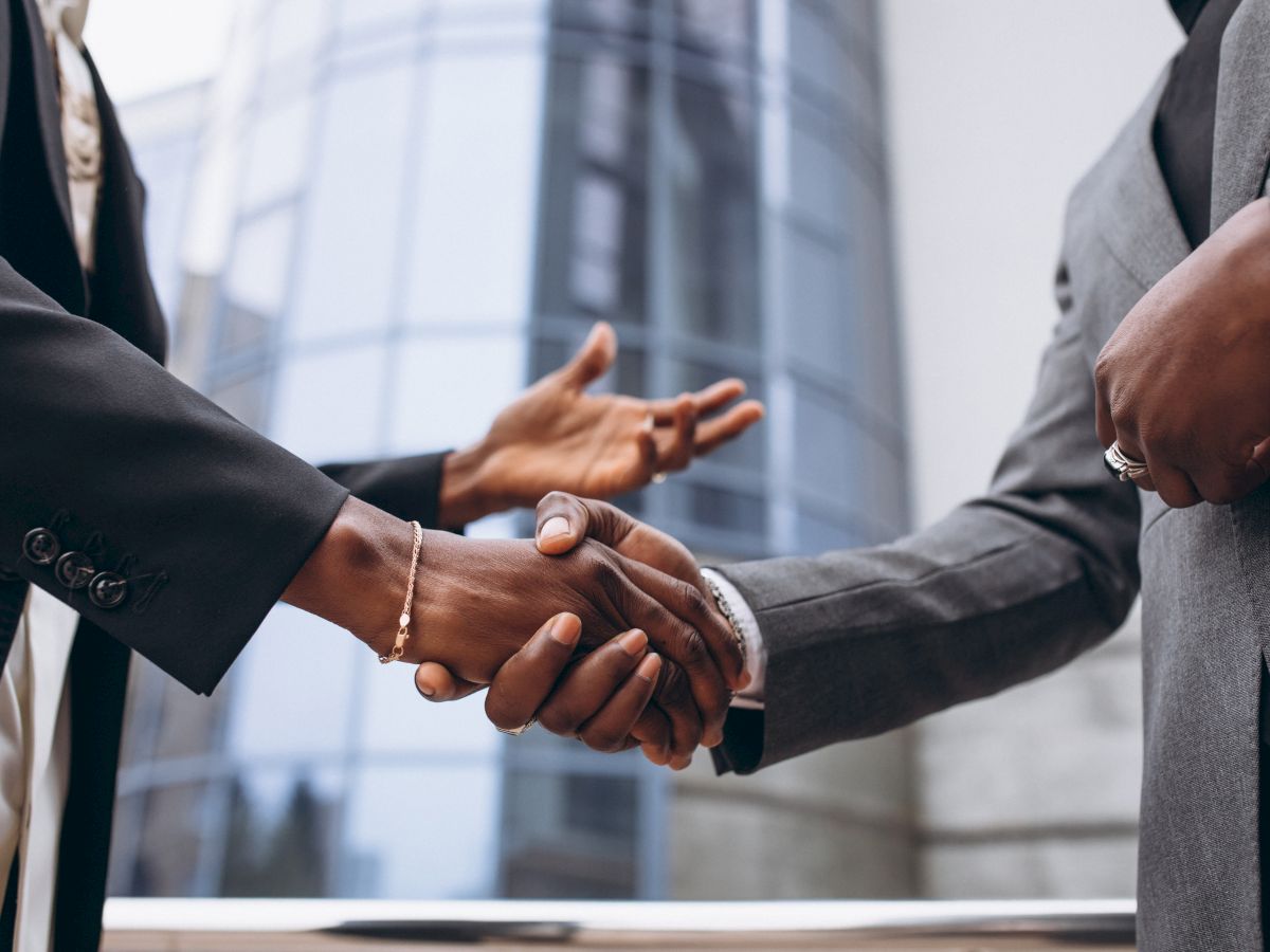 Two people in suits are shaking hands in front of a modern glass building; only their hands and arms are visible in the image.