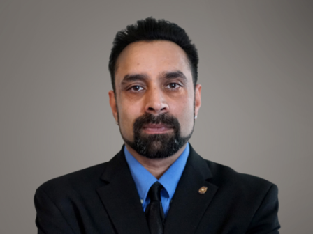 A man with a neatly trimmed beard and mustache, wearing a blue shirt and black suit jacket, poses with his arms crossed against a plain background.