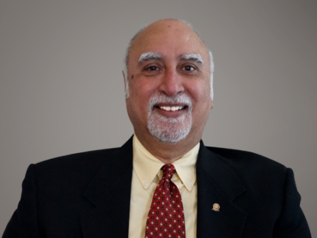 An older man with a mustache and beard smiles, wearing a suit, yellow shirt, and red tie with a dotted pattern, in front of a plain grey background.