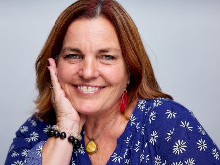 A woman with red hair smiles while resting her chin on her hand, dressed in a blue top with white floral patterns, wearing a bracelet and a necklace.