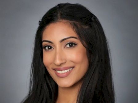 A smiling woman with long dark hair poses against a gray background, wearing a black top.