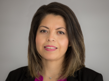 A woman with shoulder-length dark hair, wearing a black blazer over a magenta top, is looking at the camera against a plain background.