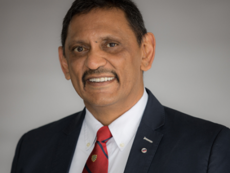 A man wearing a dark suit, white shirt, and a red patterned tie, smiling against a gray background.