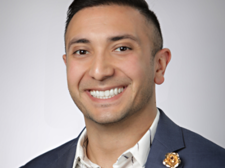 A smiling person wearing a suit and white shirt, with a decorative pin on their lapel, standing against a plain background.