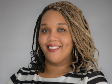 The image features a smiling person with braided hair, wearing a black and white striped shirt, posing against a plain background.