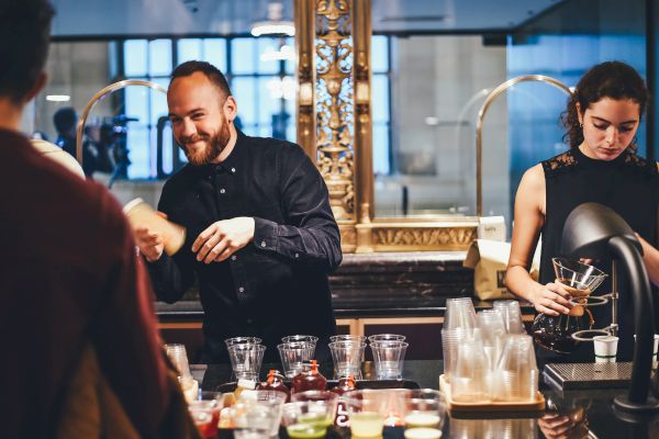 Two baristas are preparing drinks for customers behind a counter with various glasses, ingredients, and equipment in a coffee shop.