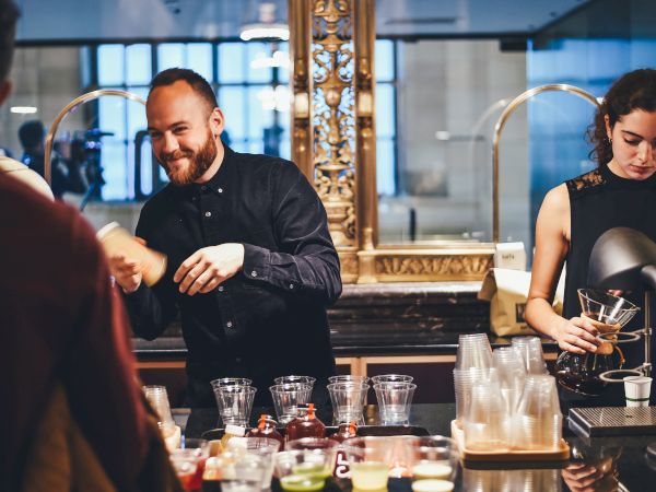 Two baristas are preparing drinks for customers behind a counter with various glasses, ingredients, and equipment in a coffee shop.