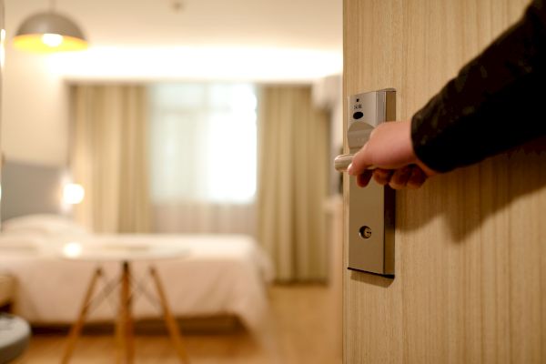 A hand is opening a door, revealing a blurred hotel room with a bed, a table, and curtains over a window, under warm lighting.