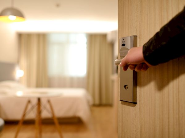 A hand is opening a door, revealing a blurred hotel room with a bed, a table, and curtains over a window, under warm lighting.