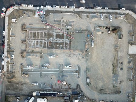 An aerial view of a construction site shows the foundational layout, various construction vehicles, and equipment scattered around the area.