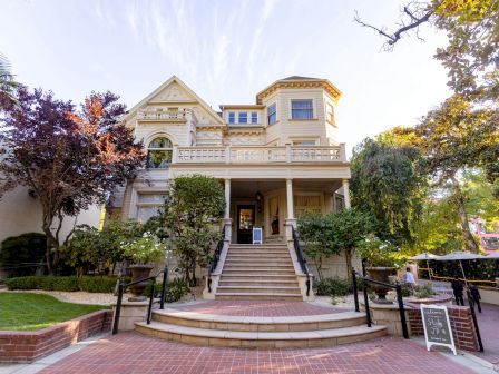 A two-story Victorian-style house with a large staircase leading to the entrance, surrounded by trees and greenery, and a signboard in front.