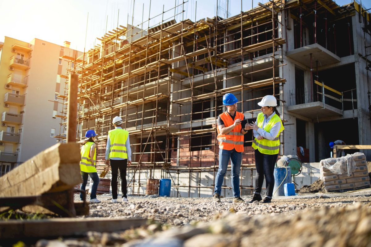 The image shows a construction site with workers wearing safety vests and helmets, discussing and inspecting the ongoing building work.